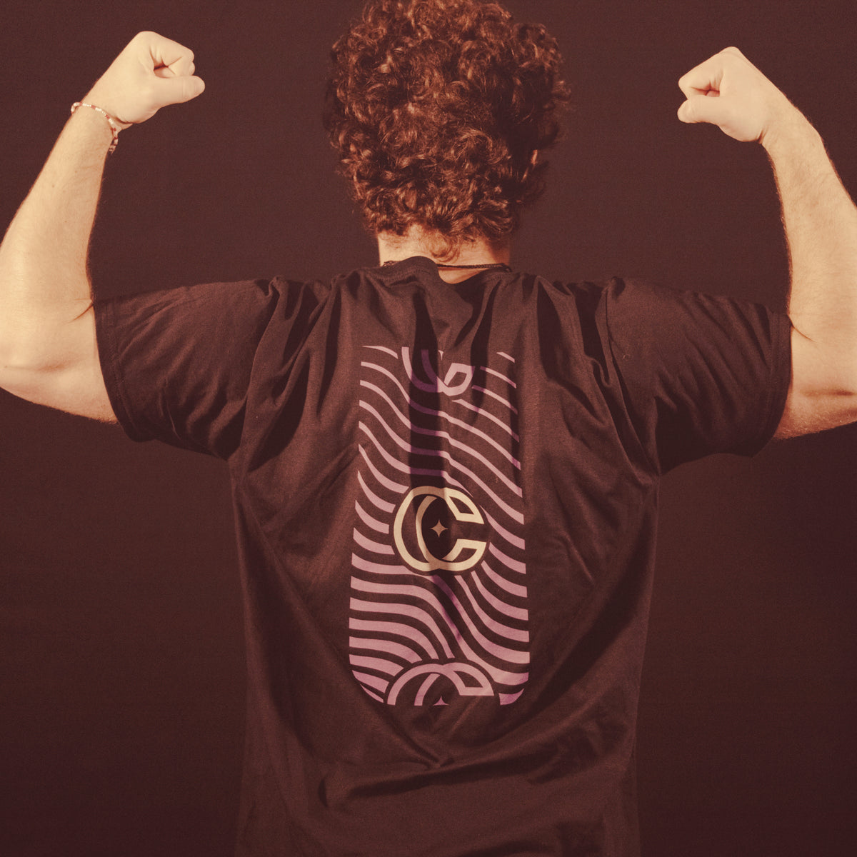 Image of a guy facing away from the camera doing a flex pose against a dark background. The back of the black shirt they are wearing has a vertical rectangle- the inside of it has purple wavy lines. the center of the rectangle has a concrete castles logo of a CC that overlaps slightly and has a star in the center. The top and bottom of the rectangle has the cc logo again, but slightly cut off and in purple.