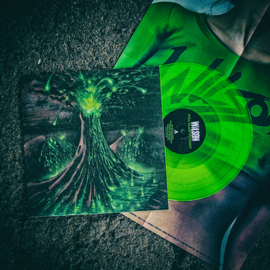 Image of the wilson- full blast fuckery album artwork on a vinyl sleeve with the lime green vinyl sticking out of the sleeve. they are lying on a grey concrete background. the album cover features a green and dark red image of a volcano erupting with green light and lava. There are red and black clouds in the sky.