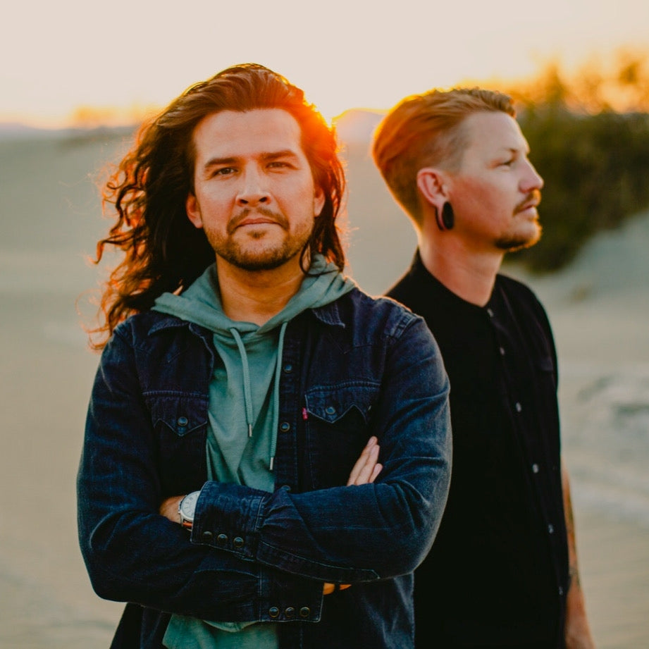 Two men stand outdoors at sunset, one with long hair and a denim jacket facing the camera with arms crossed, the other with short hair and gauged ears looking to the side.