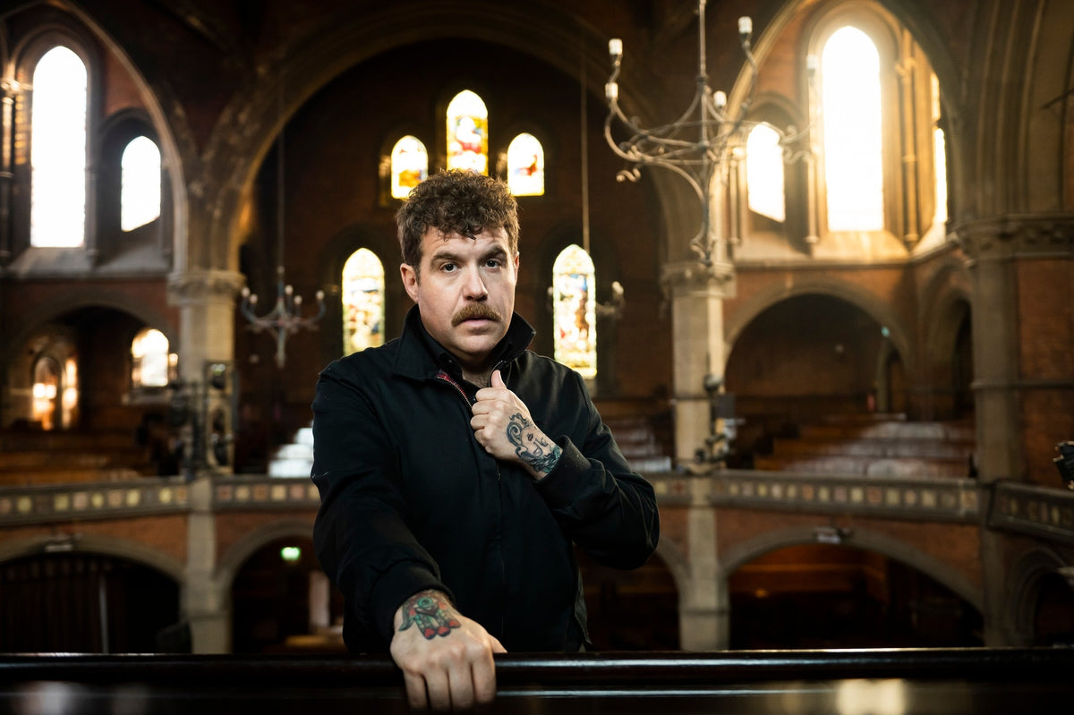A man with curly hair and a mustache adjusts his shirt collar while standing in a church with stained glass windows and arched architecture. Sunlight streams through the windows behind him.