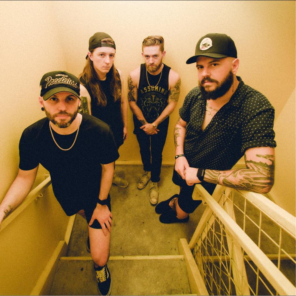 Four men with tattoos and casual clothing stand on a staircase in a narrow, yellow-lit hallway, looking up at the camera with serious expressions.