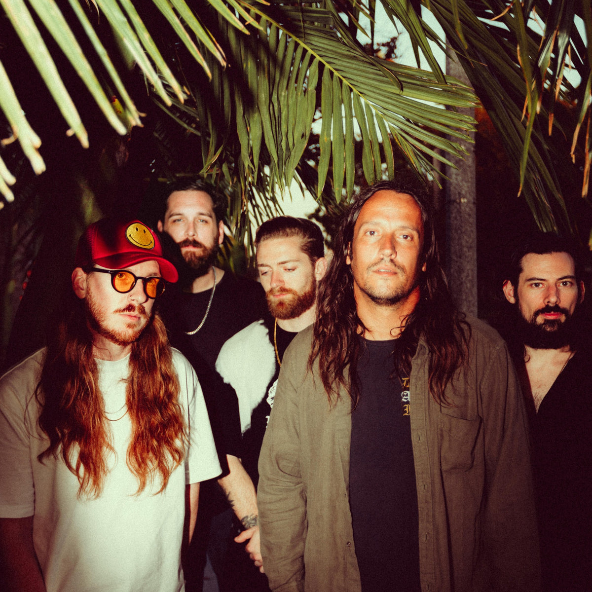 Five men with long hair and beards stand under palm leaves. One wears a red cap with a smiley face and tinted glasses; others wear casual dark clothing. The background is dimly lit and tropical.