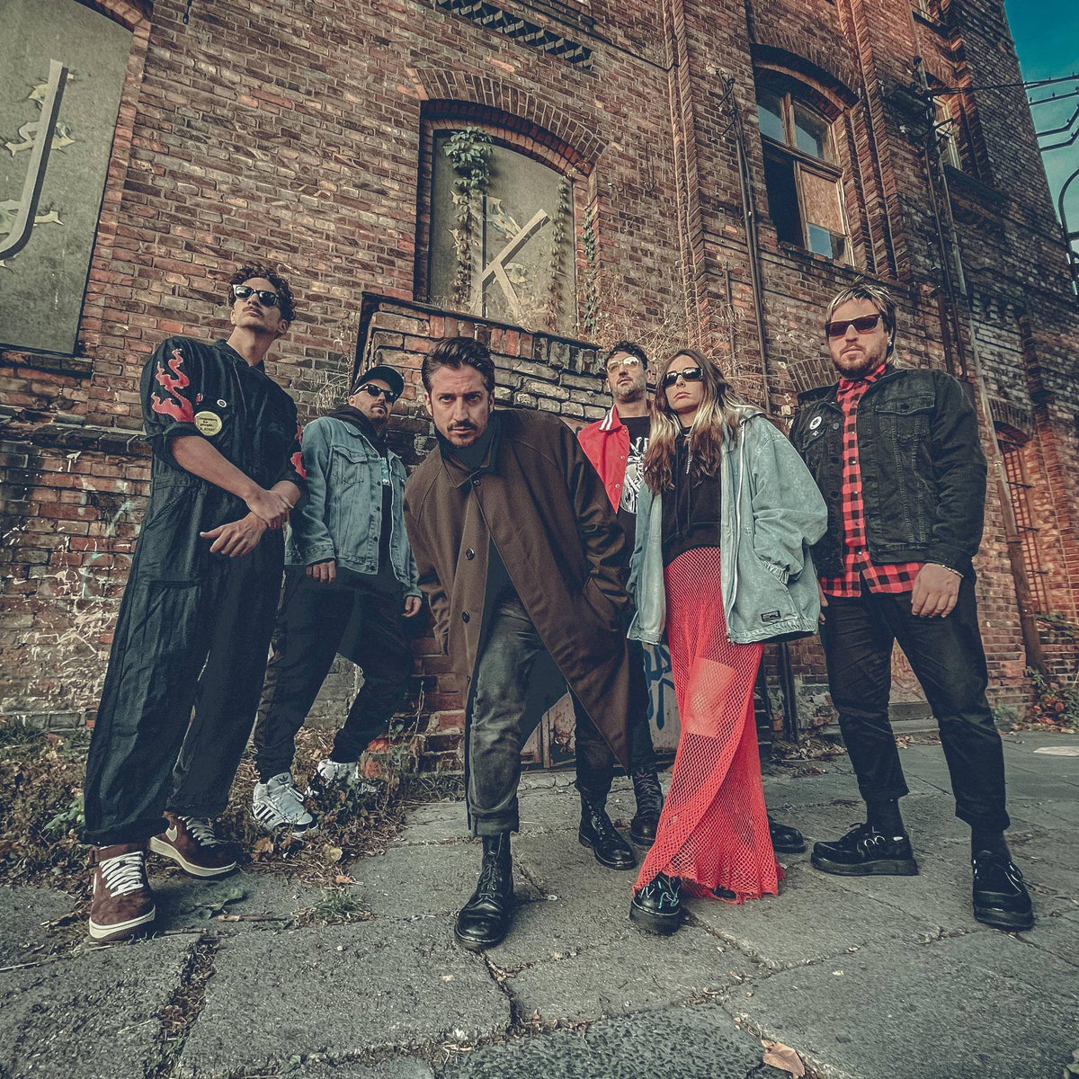Six people pose confidently in urban streetwear in front of an old brick building with tall windows, showing a mix of serious and cool expressions. The atmosphere is edgy and stylish.
