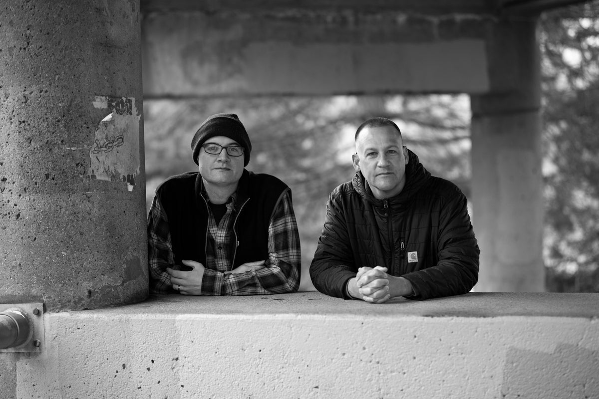Two men stand side by side at a concrete railing outdoors. One wears glasses, a beanie, and a plaid shirt, while the other wears a dark jacket. Both look toward the camera in black and white.