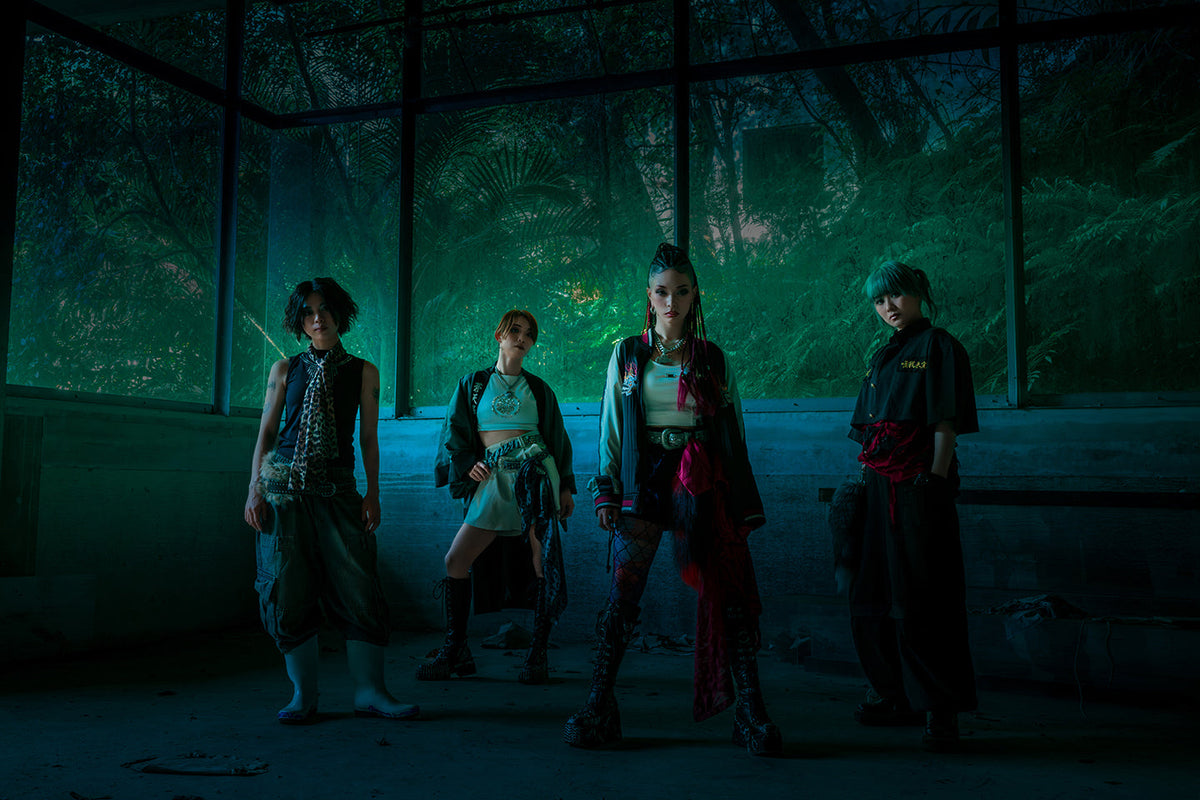 Four people in diverse, edgy outfits stand in a line in a dimly lit, abandoned space with large windows looking out onto a dark, lush forest.