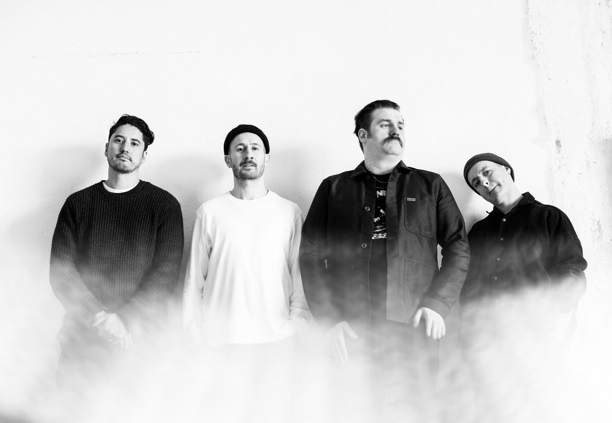 Four men standing in a line against a white wall with fog at their feet.