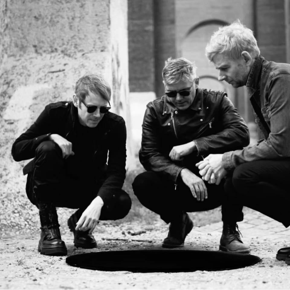 Three men in dark clothing crouch around a large, circular hole in the ground, looking inside with serious expressions. The photo is in black and white.