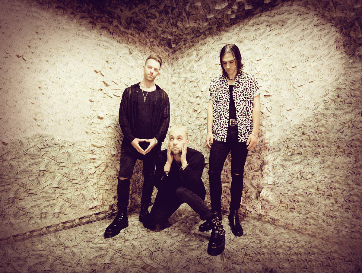 Three people pose in a room covered with dollar bills. One stands with hands making a heart, one crouches with hands on their face, and another stands with hands in pockets, all wearing dark, stylish clothing.