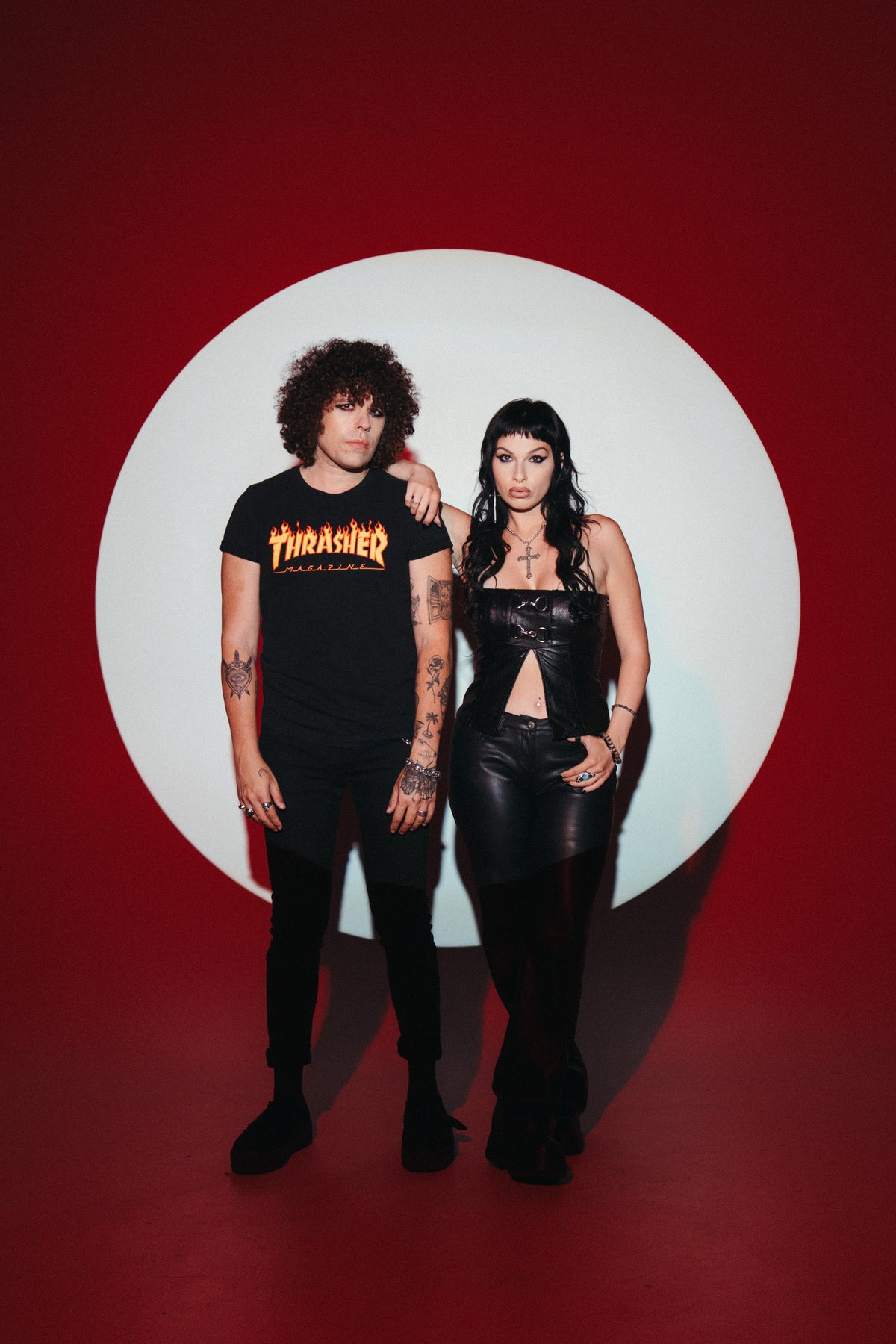 A man in a Thrasher t-shirt and a woman in a black top and pants stand in front of a large white circle on a red background.