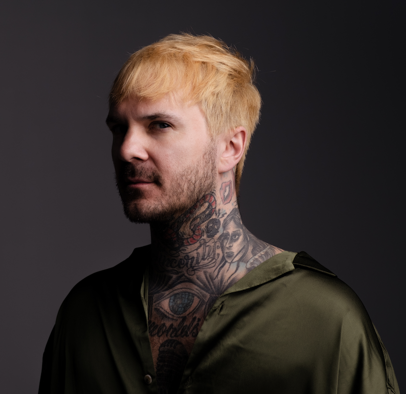 A person with short blond hair, facial hair, and extensive neck and chest tattoos wears an olive green shirt against a dark, gradient background.