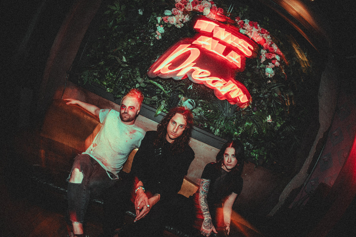Three people sit on a bench in front of a neon sign that reads Takes All Dream, surrounded by flowers and greenery, in a dimly lit, moody room.