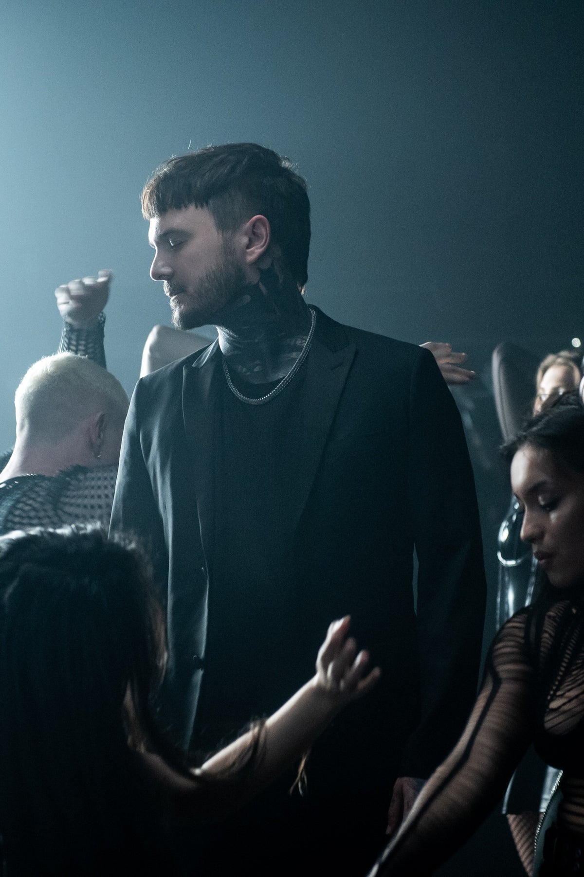 A man in a black suit with neck tattoos stands among people dancing in a dimly lit, smoky room. He looks to the side while others raise their arms and dance around him.