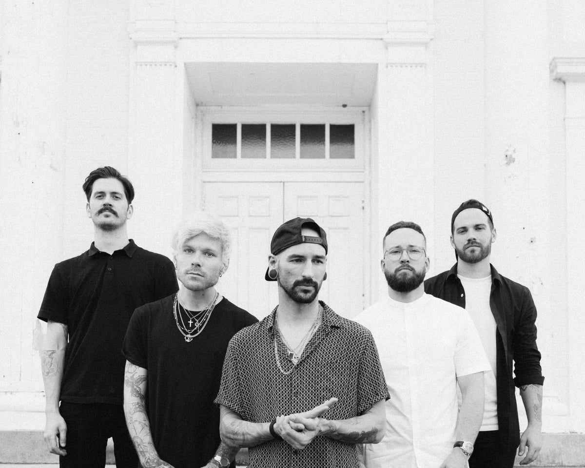Five men with tattoos and facial hair stand in front of a white building, posing for a group photo. They wear casual, dark clothing and have serious expressions.
