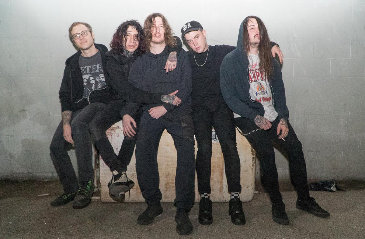 Five men with long hair and tattoos sit closely together on a concrete block against a plain wall, looking relaxed. One holds a cigarette, and they are dressed in casual, dark clothing.
