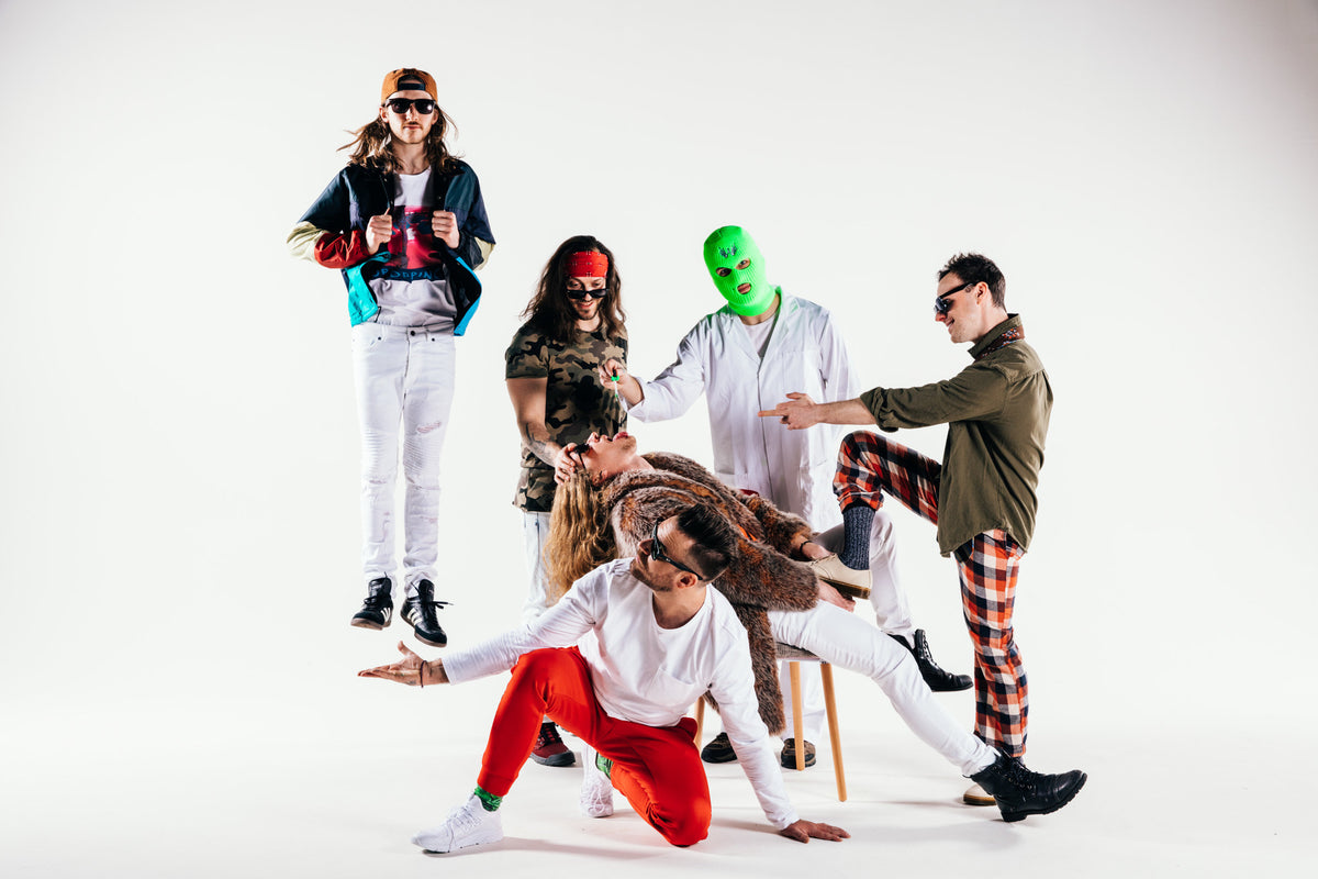 Six people in quirky costumes pose playfully; one floats mid-air, another wears a green mask, and others strike dramatic, energetic stances against a plain white background.