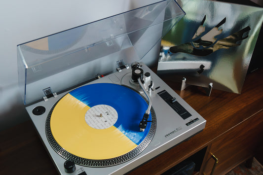 A Limited Edition Vinyl by Thursday, with yellow and blue halves, spins on a silver turntable with a clear dust cover; the abstract art album cover rests nearby on a wooden surface.