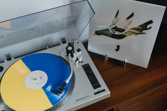 A Velocity Records exclusive: a Thursday Waiting vinyl, half blue and half yellow, spins on a silver turntable, with its abstract art sleeve nearby on a wooden surface.