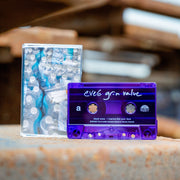 Cassette tape and cassette case against a blurred brown and white background. The tape is a clear blue and says eve 6 grim value in white text. the cassette case is an image of four cases of bottled water, with cigarettes in a clear plastic baggie sitting on top of one of the cases of water.
