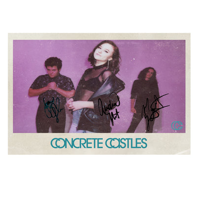Image of a white postcard with a pink colored photo in the center. the pink colored photo has the 3 members of concrete casteles in it, and they have all signed their names near themselves on the postcard. there is a female standing sideways, looking at the camera wearing black. behind here are two males wearing black and looking at the camera. below this image in blue text reads "concrete castles".
