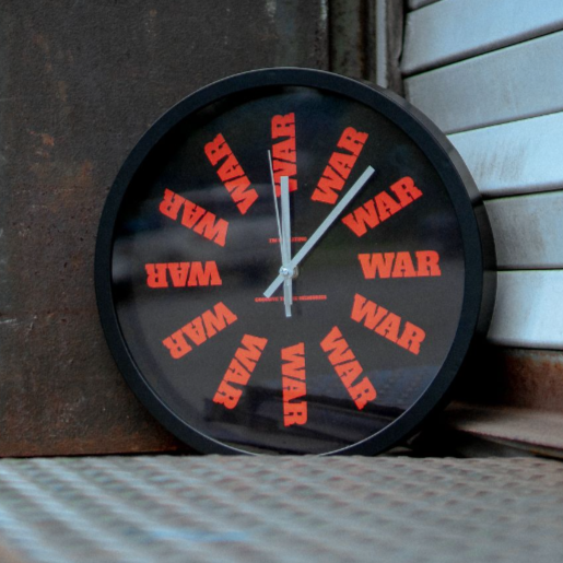 Black wall clock with war written in red in place of numbers. The hands of the clock are white. The clock is leaning against a rusted wall and on top of a metal surface.