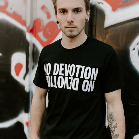 A person with short hair and ear gauges stands by a graffiti wall, wearing a black No Devotion t-shirt with the brand name printed upside down in white letters.