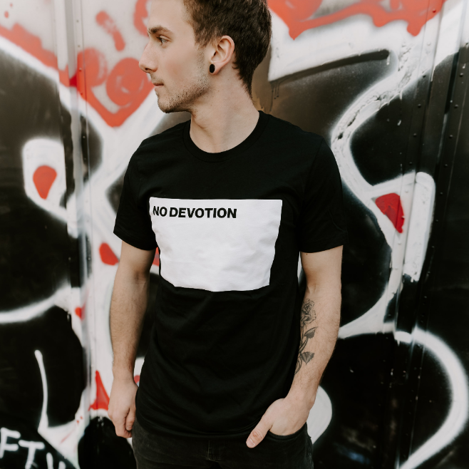 A man with short hair and earrings, wearing a black No Devotion T-shirt, stands before bold black, red, and white graffiti, looking to his left.