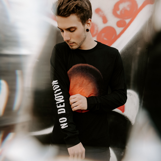 A person with short hair and ear gauges adjusts the sleeve of their black No Devotion shirt, which features NO DEVOTION text on the arm and an orange-red circular design on the chest, standing against a graffiti wall.