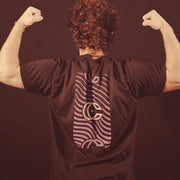 Image of a guy facing away from the camera doing a flex pose against a dark background. The back of the black shirt they are wearing  has a vertical rectangle- the inside of it has purple wavy lines. the center of the rectangle has a concrete castles logo of a CC that overlaps slightly and has a star in the center. The top and bottom of the rectangle has the cc logo again, but slightly cut off and in purple.