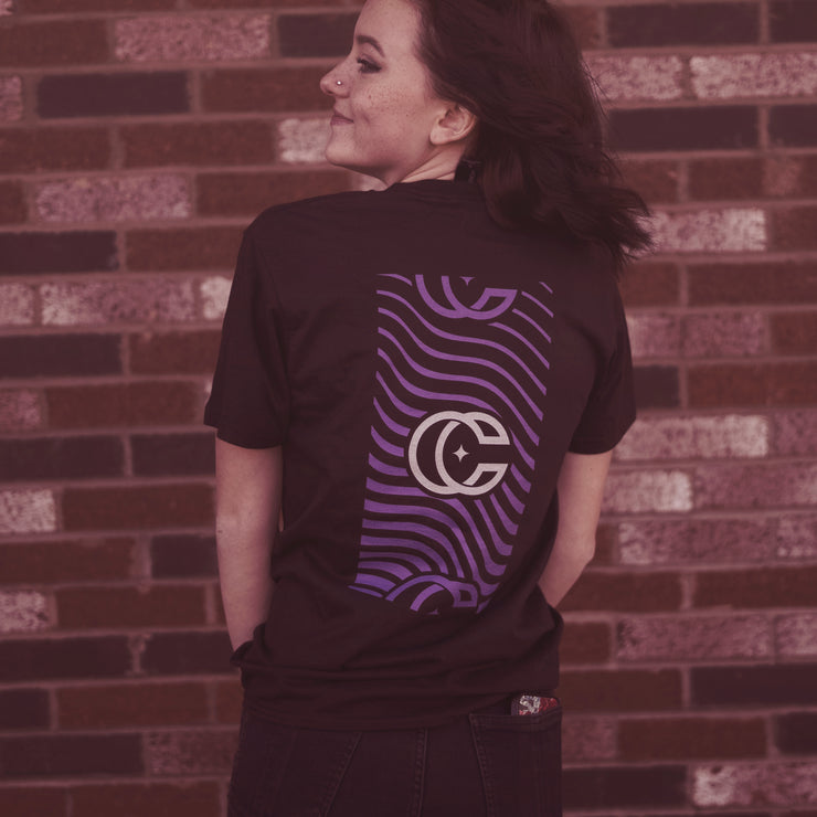 image of a girl with her back to the camera, head turned to the left. she has medium length dark hair with a brick wall in the background. the girl is wearing a black tshirt and black jeans.  the back of the shirt is a vertical rectangle- the inside of it has purple wavy lines. the center of the rectangle has a concrete castles logo of a CC that overlaps slightly and has a star in the center. The top and bottom of the rectangle has the cc logo again, but slightly cut off and in purple.