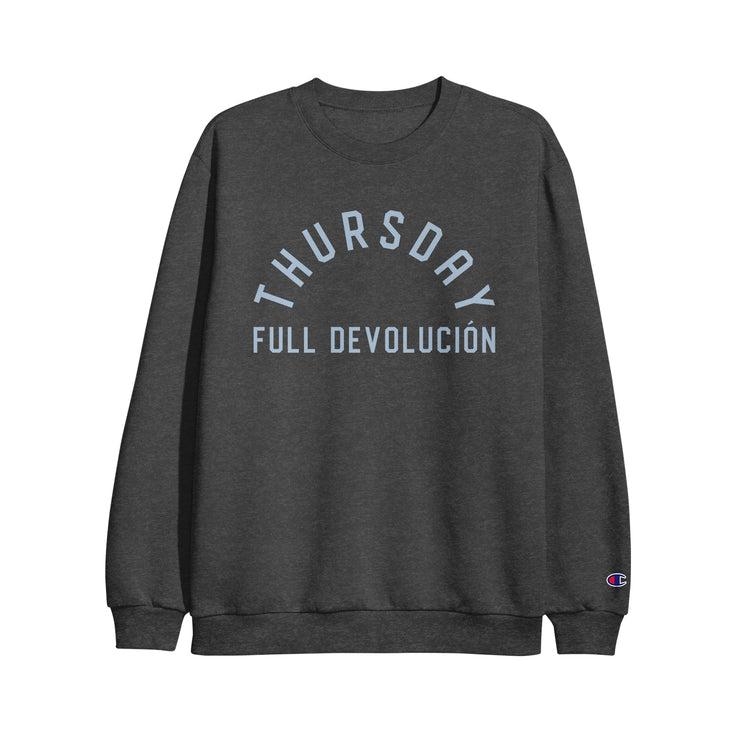 Image of a charcoal heather crewneck against a white background. Across the chest in an arch reads "thursday". Below that it says "full devolucion". The writing is in a light blue color. The left sleeve at the bottom has the champion C logo in red, white and blue.