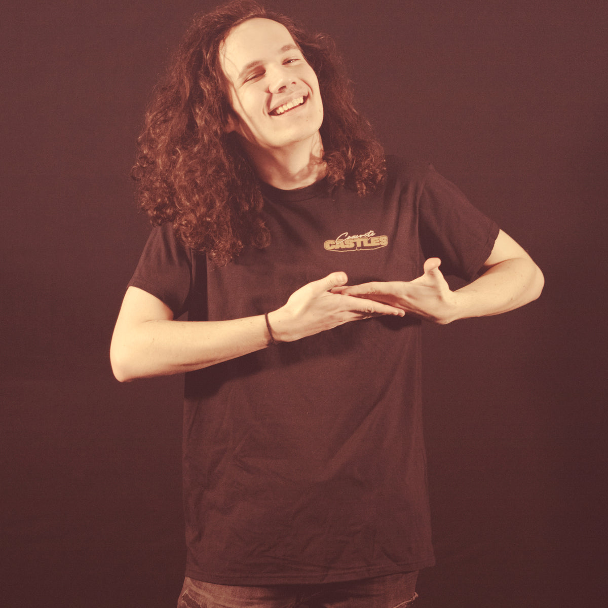 Image of a guy wearing a black tshirt, facing the camera against a dark background. they have their hands up by their chest to show off the logo on the shirt.  the left chest says concrete castles. the word concrete is in white cursive, and the word castles is in regular bold dark gold text.