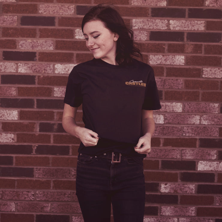 image of a girl with medium length dark hair looking to her left with a brick wall in the background. the girl is wearing a black tshirt and black jeans. the left chest of the tshirt says concrete castles. the word concrete is in white cursive, and the word castles is in regular bold dark gold text.