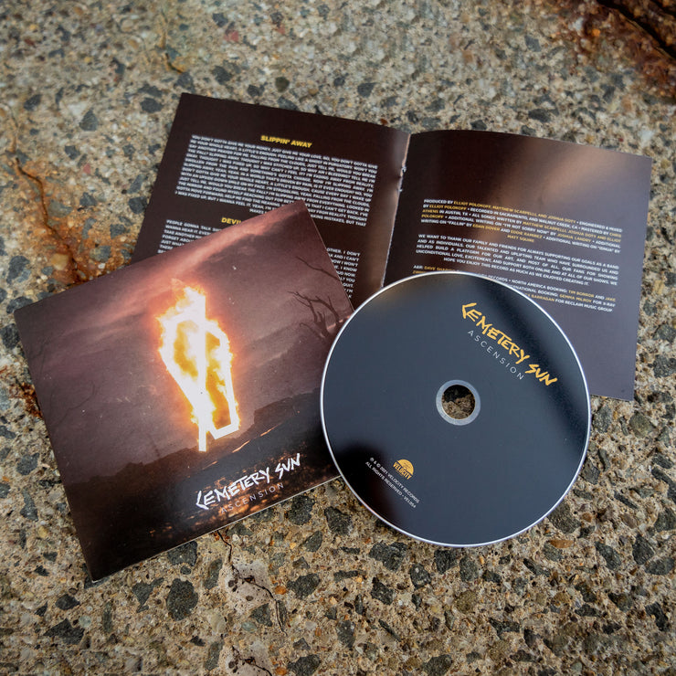  CD case, booklet, and cd against a grey concrete background. the cd cover is an Image of a burned field with a dark sky. There is a coffin shape in the center that is on fire. Below this in white text reads "cemetery sun, ascension". The cd itself is black and says cemetery sun ascension in gold and white text. The booklet is gold and white and features lyrics to the songs on the cd.